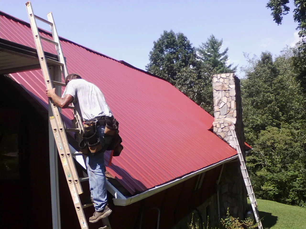 Roofer on a ladder beside a red metal roof and stone chimney — Soffit & Fascia Repair by Roofing Brothers in Tulare